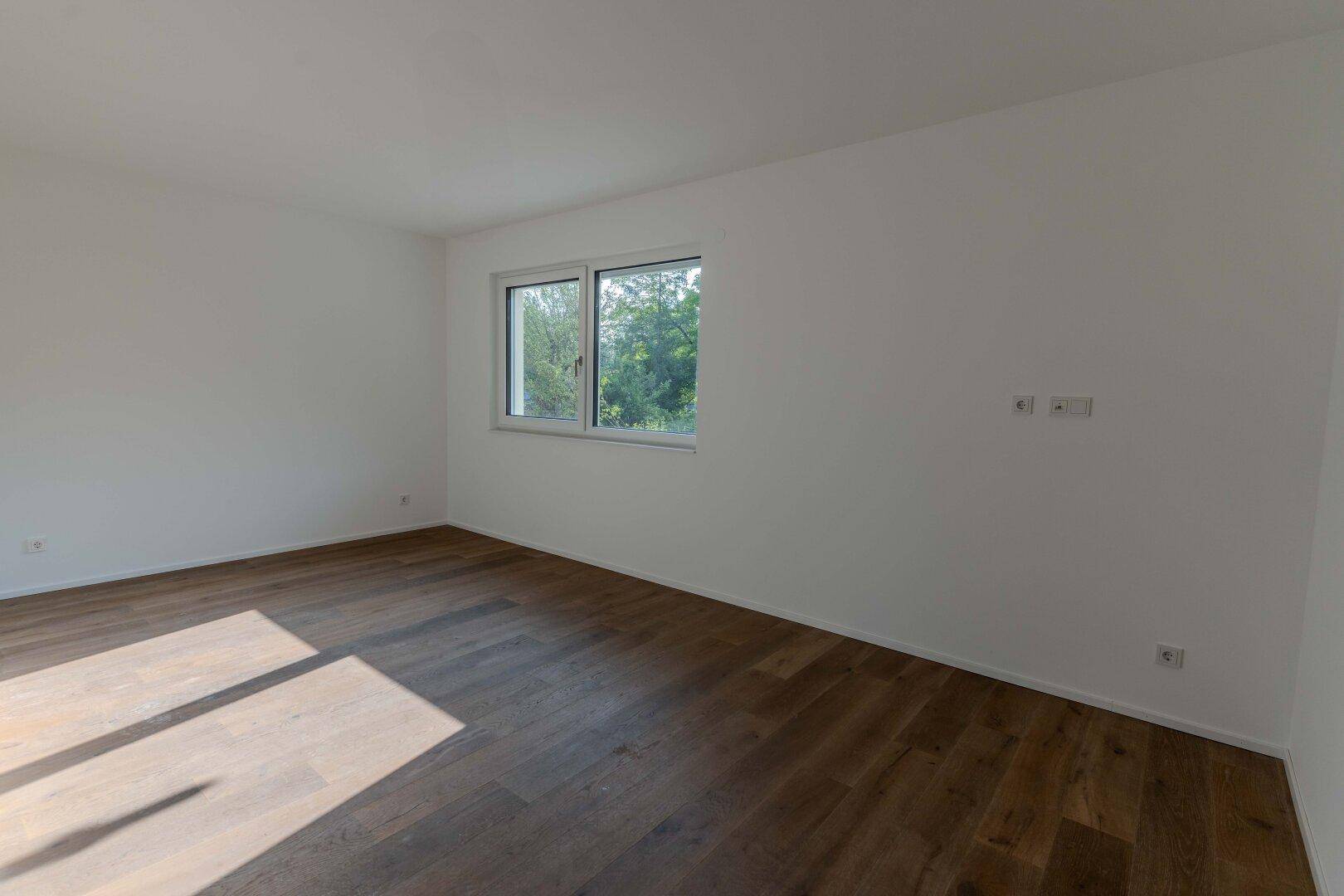 Schlichtes Zimmer mit Holzboden und einem Fenster, das viel natürliches Licht hereinlässt.