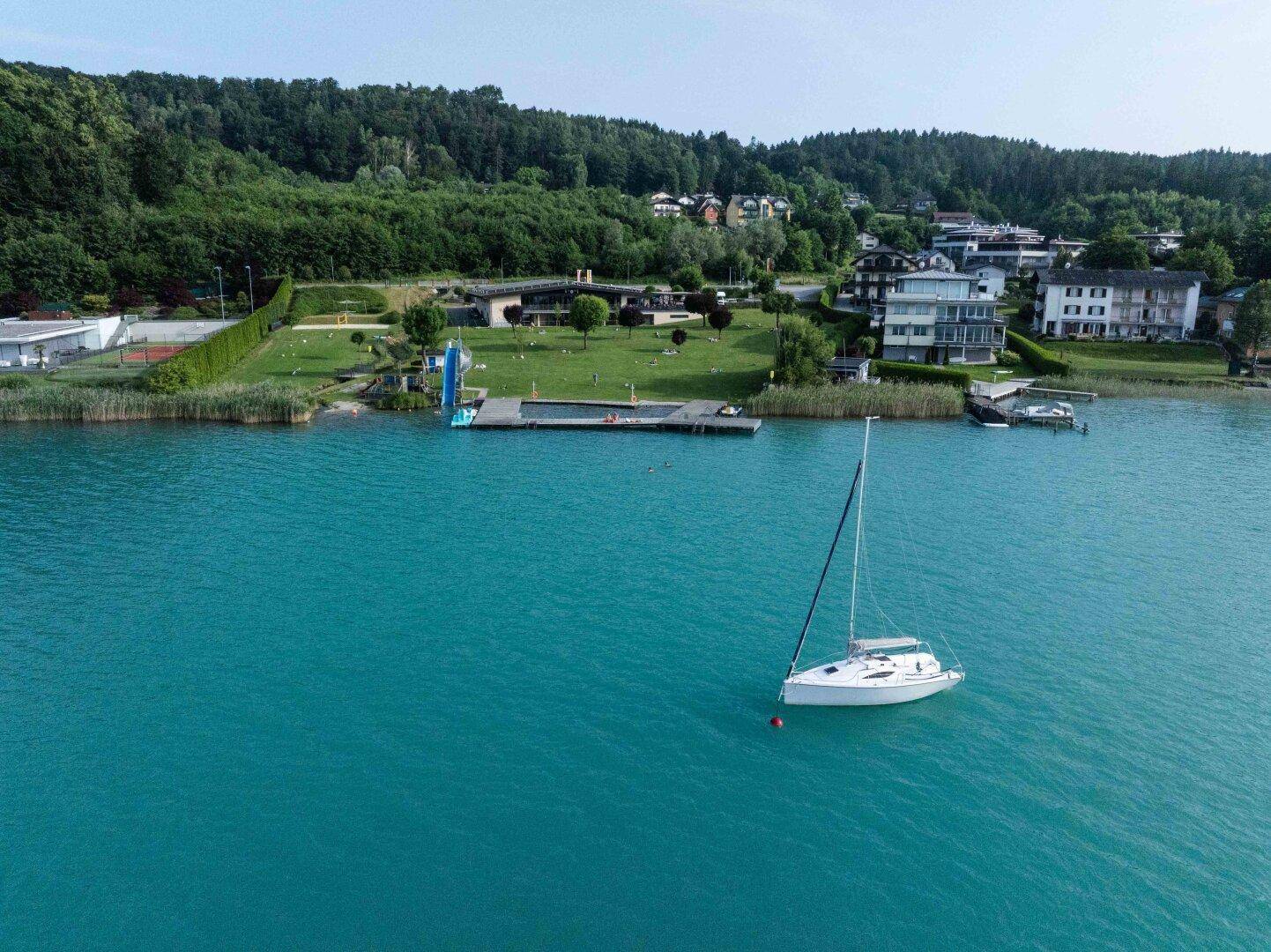 Segelboot auf einem blauen See mit Uferbebauung, Grünflächen und einem Poolbereich.