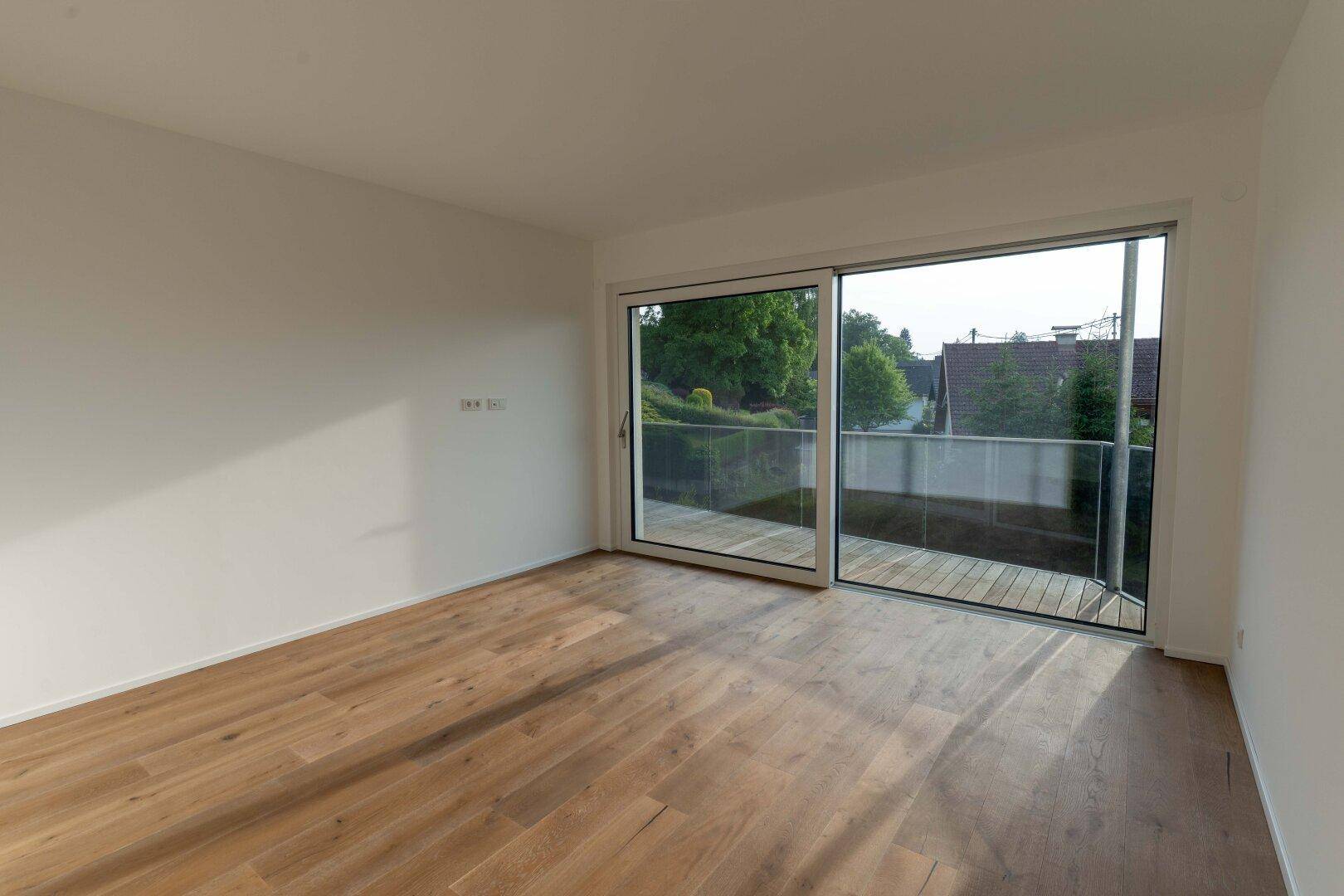 Heller Raum mit Holzdielenboden und großer Schiebetür, die zu einem Balkon mit Ausblick führt.