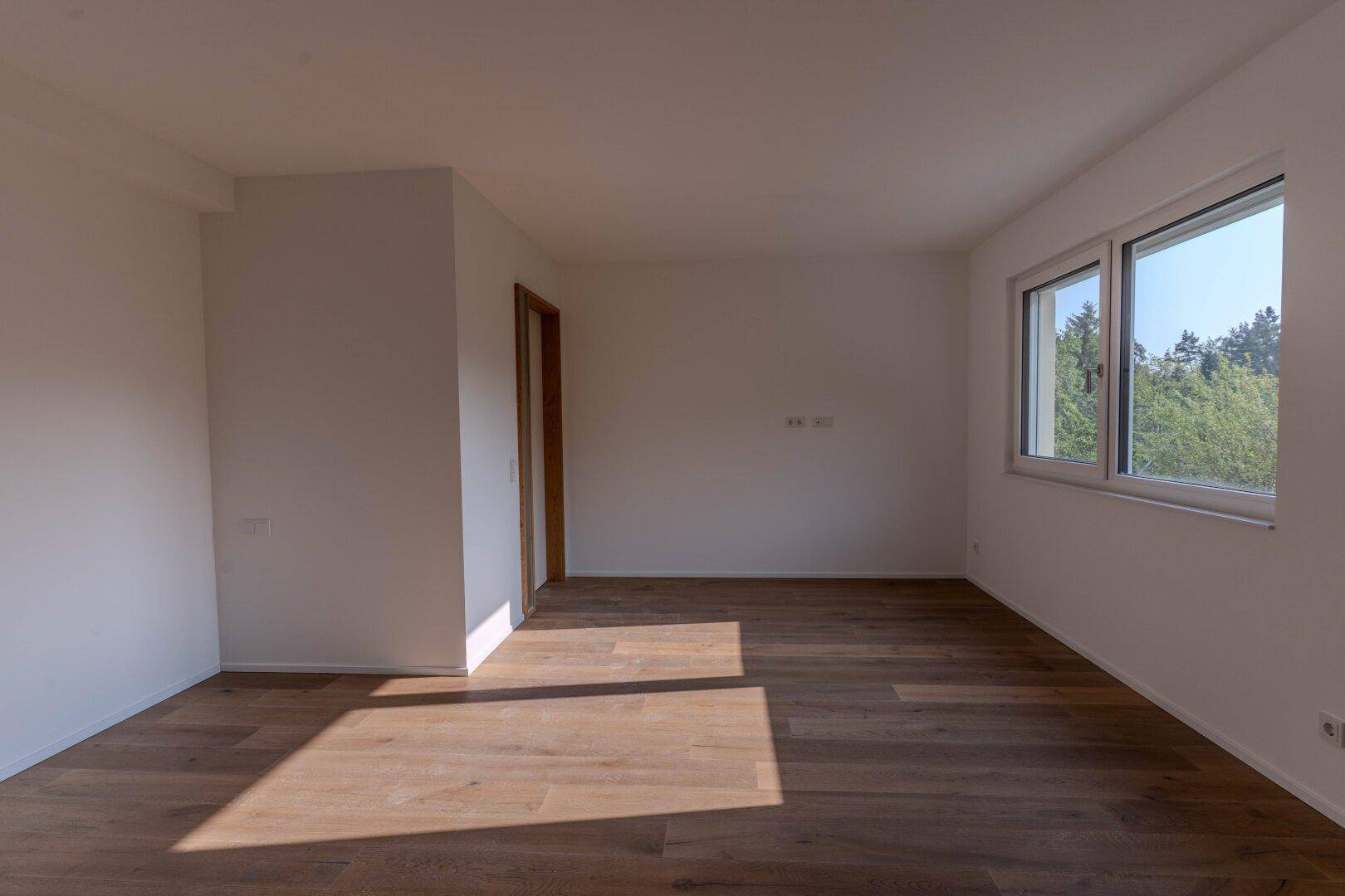 Leerer Raum mit Holzdielenboden und zwei Fenstern, die einen Blick ins Grüne bieten.