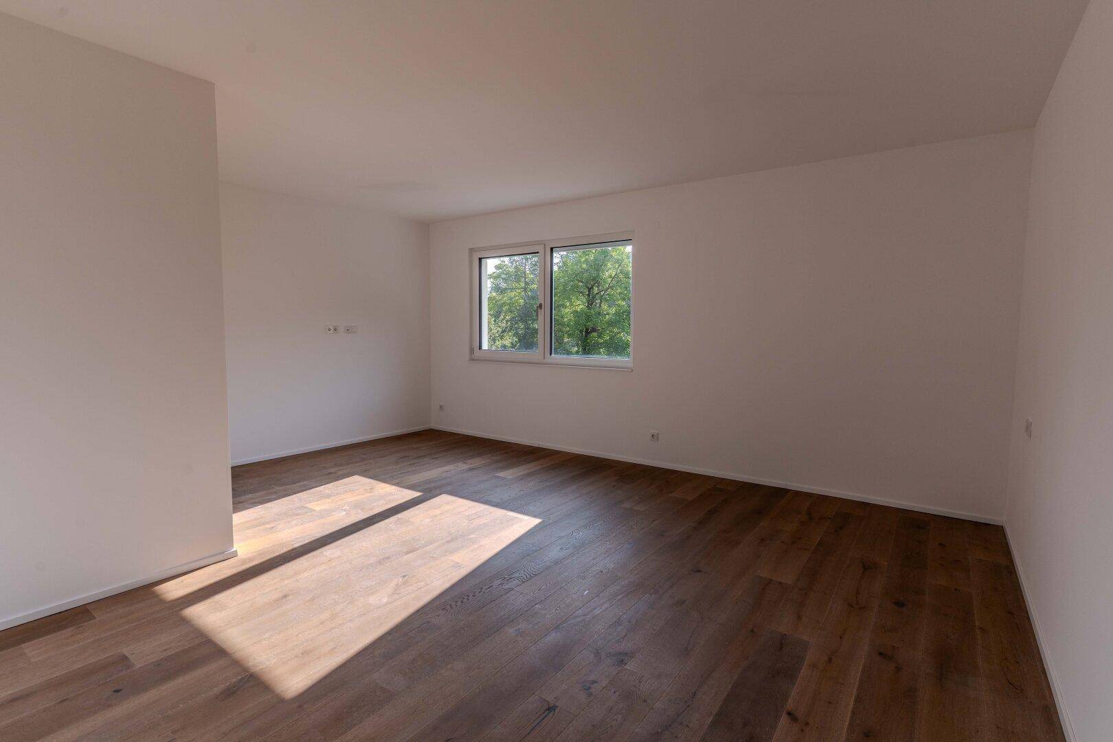 Leerer Raum mit Holzdielenboden und zwei Fenstern, die einen Blick ins Grüne bieten.
