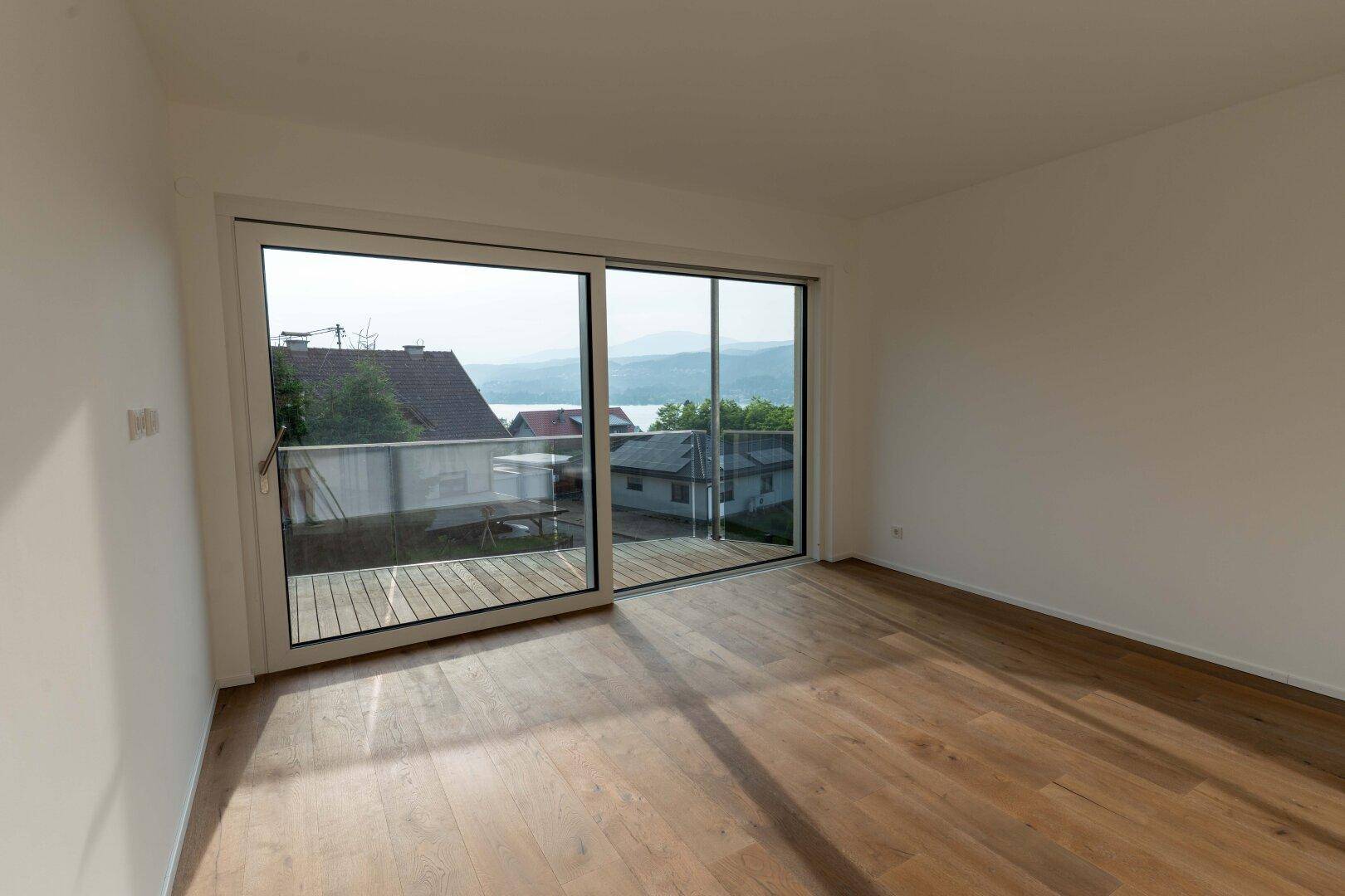 Heller Raum mit Holzdielenboden und großer Schiebetür, die zu einem Balkon mit Seeblick führt.