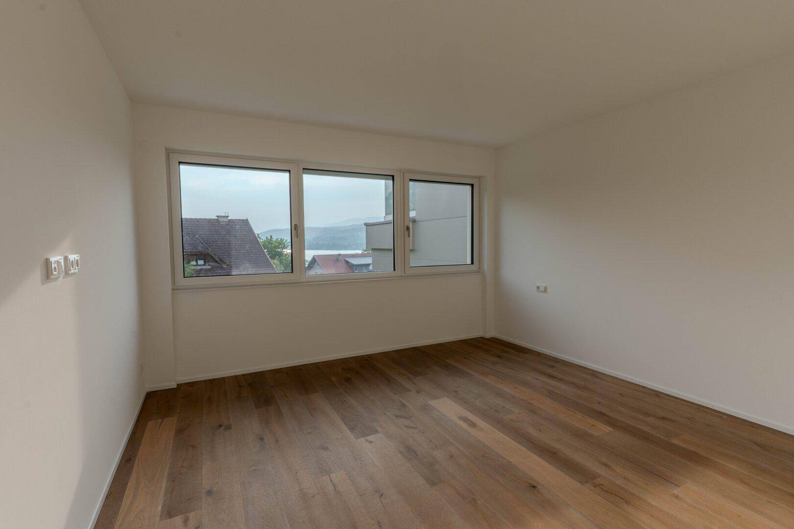 Heller Raum mit Holzdielenboden und drei Fenstern, die einen weiten Blick auf den See ermöglichen.