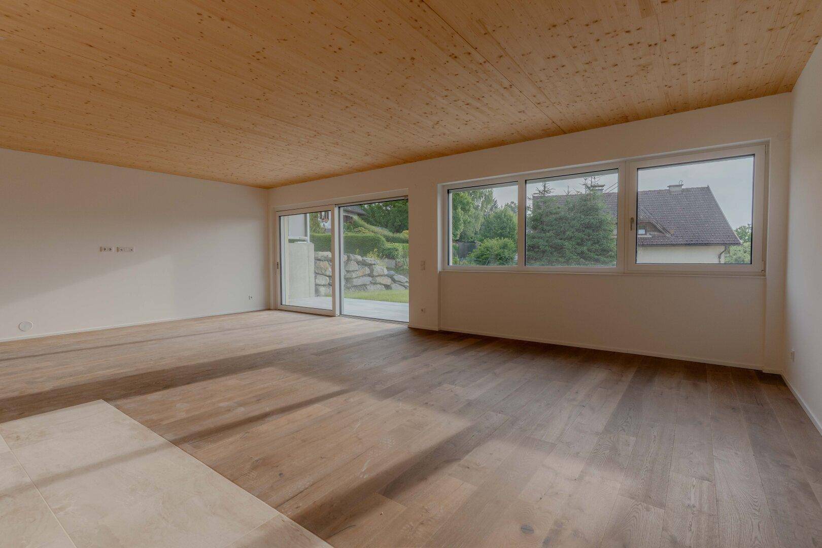 Heller Wohnraum mit Holzdielenboden, Holzdecke und großen Fenstern mit Zugang zum Garten.