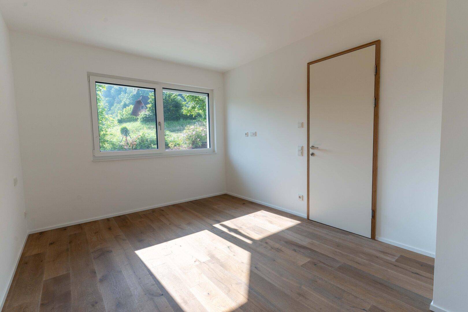 Helles Schlafzimmer mit Holzboden und Fenster, das einen Blick ins Grüne bietet.