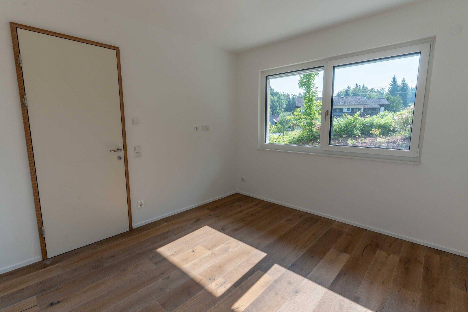 Helles Schlafzimmer mit Holzboden und Fenster, das einen Blick ins Grüne bietet.