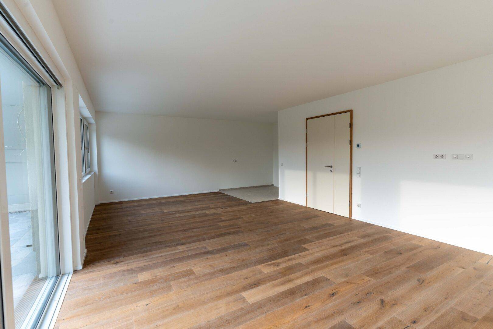Geräumiger, unmöblierter Wohnbereich mit Holzboden und Zugang zu einer Terrasse.