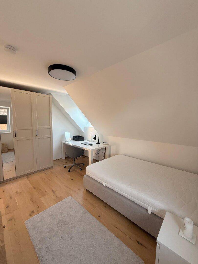 Helles Schlafzimmer mit Einzelbett, Schreibtisch und Kleiderschrank unter der Dachschräge.