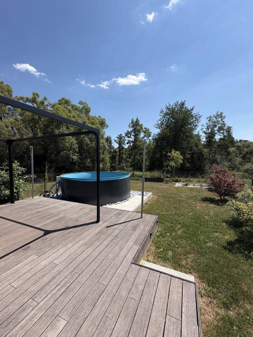 Holzterrasse mit kleinem Pool und Blick ins Grüne unter blauem Himmel.
