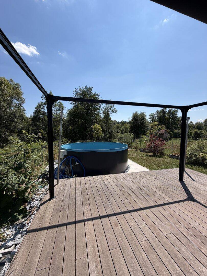 Sonnige Holzterrasse mit Pool und Pergola-Struktur, umgeben von Natur.