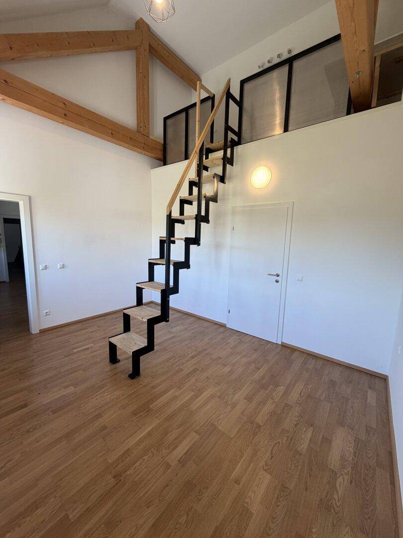 Heller Wohnraum mit offener Treppe zu einem Loftbereich und sichtbaren Holzbalken.