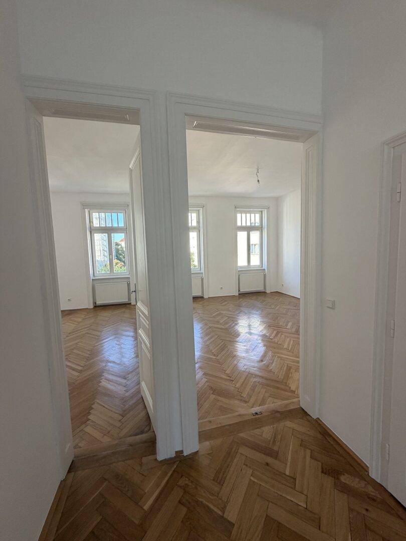 Zwei helle, leere Zimmer mit Fischgrätparkettboden, verbunden durch eine breite Türöffnung.