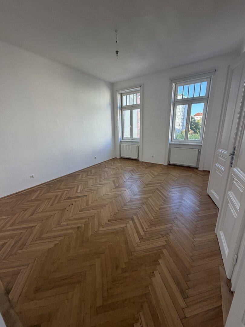 Geräumiges, leeres Zimmer mit Fischgrätparkettboden und zwei großen Fenstern, ideal für Wohnzwecke.
