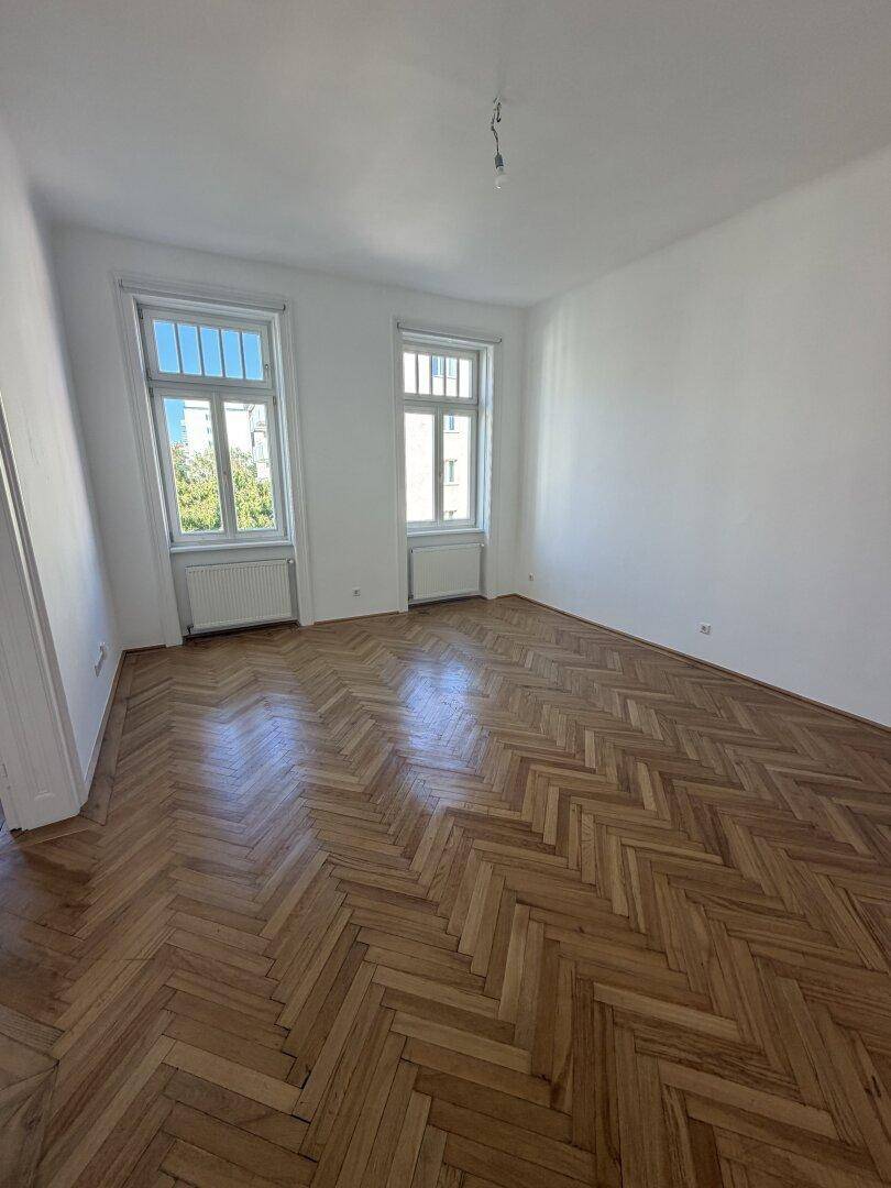 Leeres Zimmer mit glänzendem Fischgrätparkettboden und zwei Fenstern, das viel Tageslicht hereinlässt.