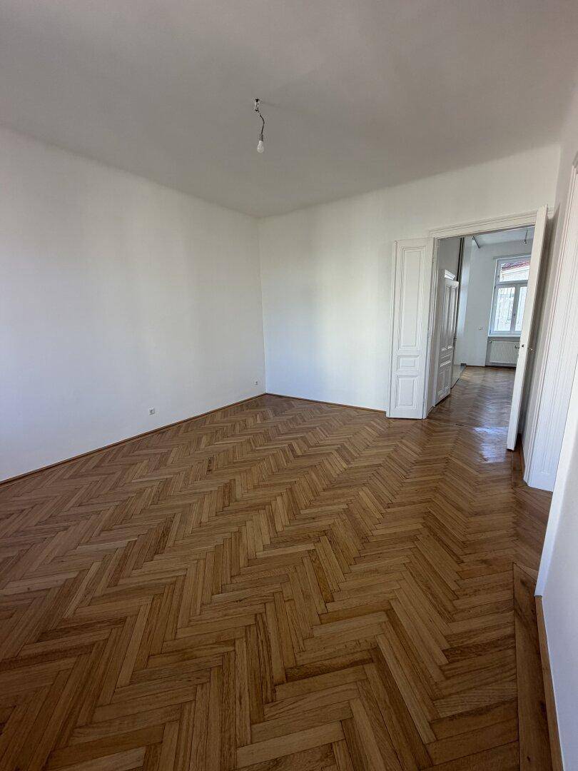 Geräumiges, leeres Zimmer mit Fischgrätparkettboden und Zugang zu einem weiteren Raum.