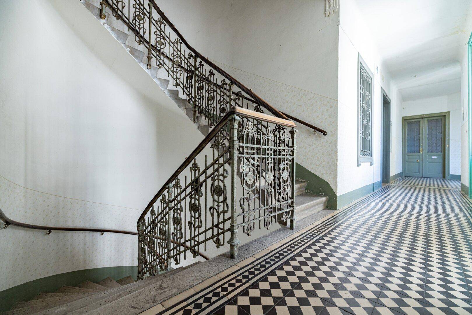 Historisches Treppenhaus mit kunstvollem Geländer und schwarz-weißem Fliesenboden im Altbau.