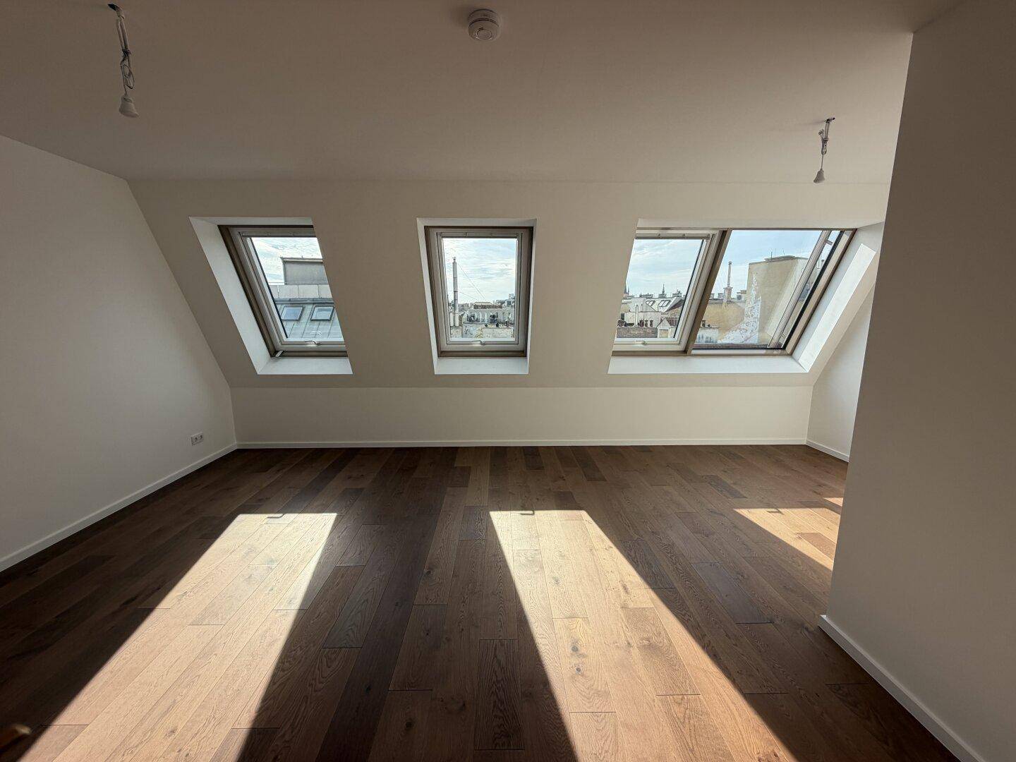 Geräumiges, unmöbliertes Zimmer im Dachgeschoss mit Holzboden und drei Dachfenstern.