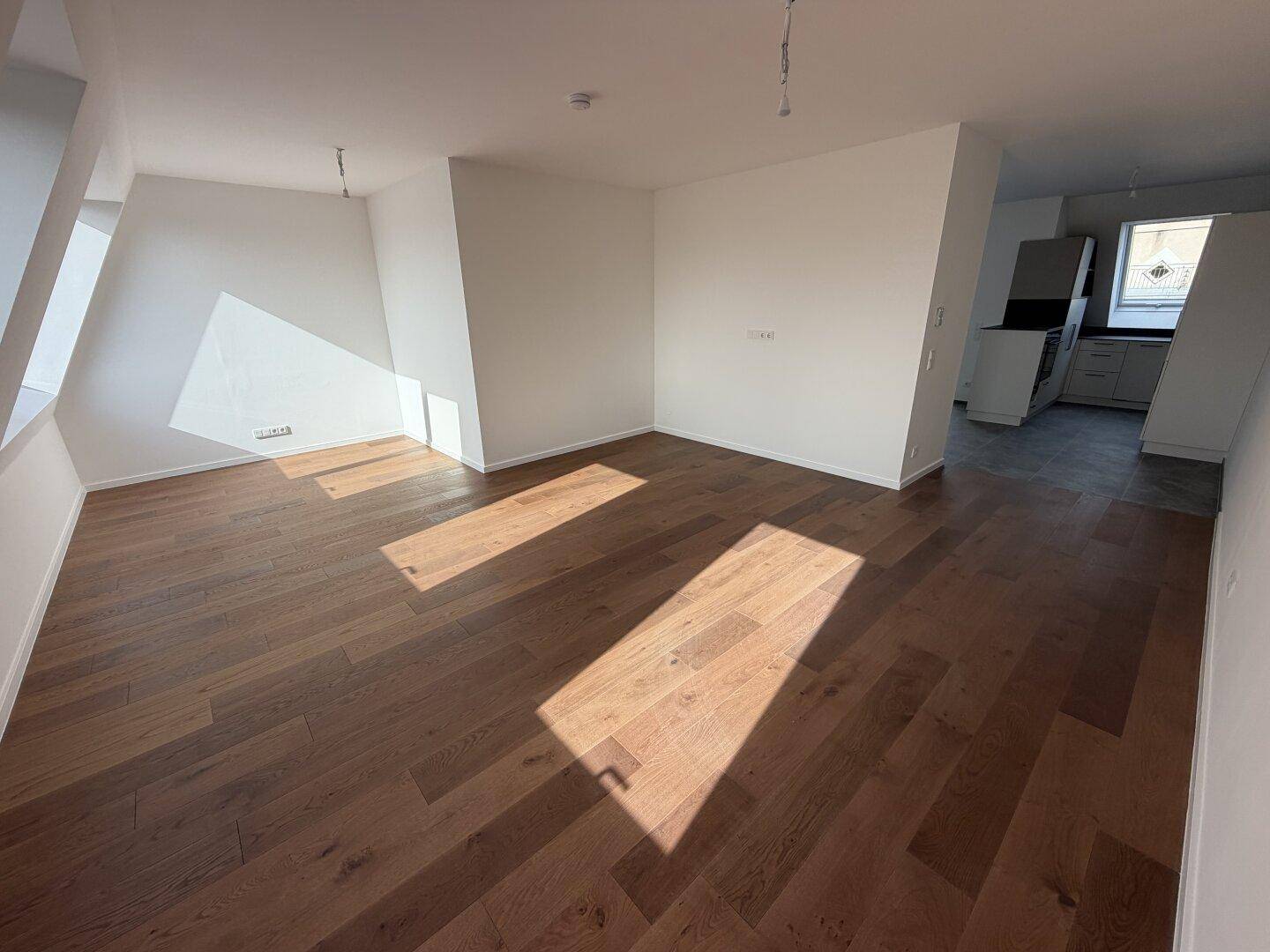 Geräumiges, unmöbliertes Zimmer mit Holzboden und Blick auf eine moderne Küche.