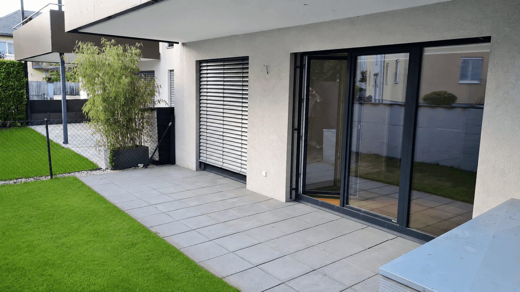 Moderne Terrasse mit Steinplatten und angrenzender Rasenfläche, zugänglich durch große Glastüren.