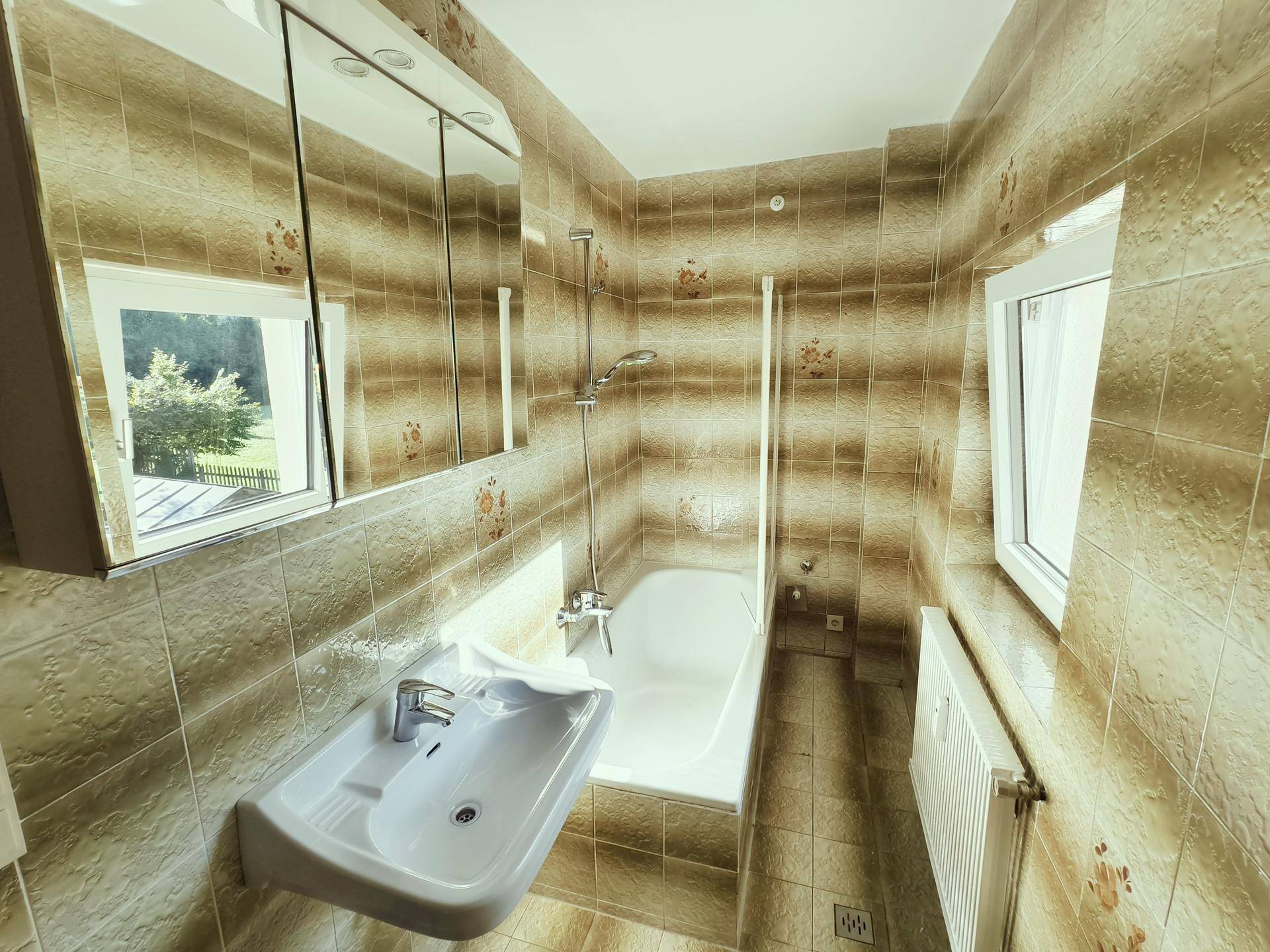 Gefliestes Badezimmer mit Badewanne, Waschbecken und Fenster mit Blick ins Grüne.