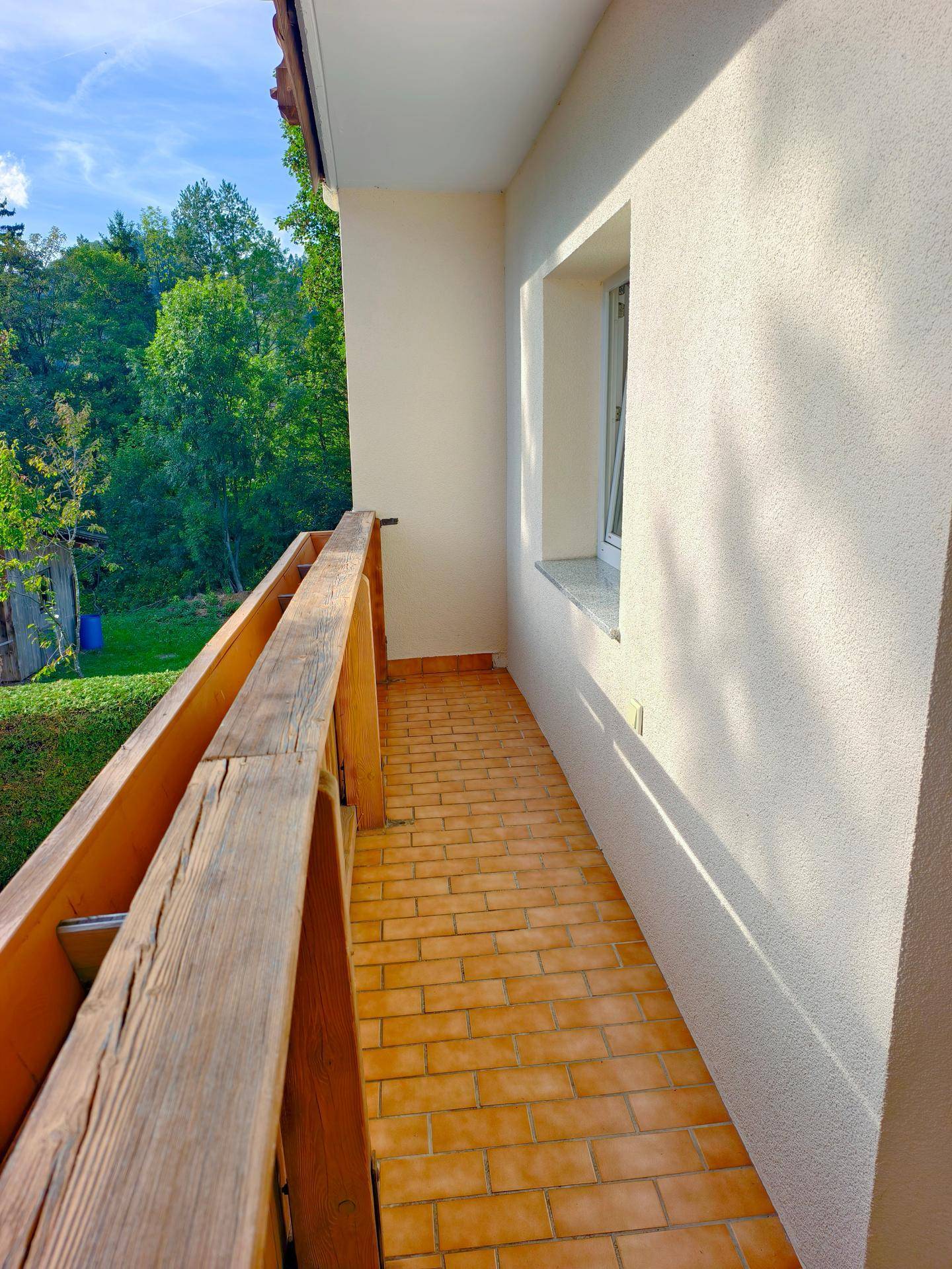 Schmaler Balkon mit Ziegelboden und Holzgeländer, umgeben von grüner Natur.