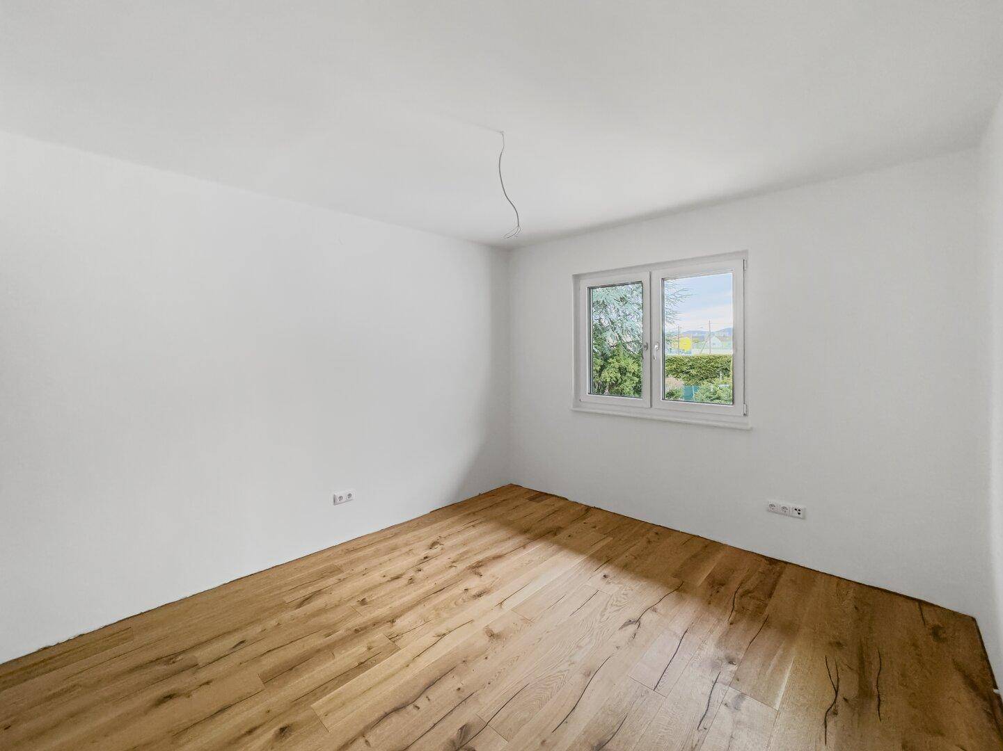 Helles, unmöbliertes Schlafzimmer mit Holzboden und Fenster, das einen grünen Ausblick bietet.