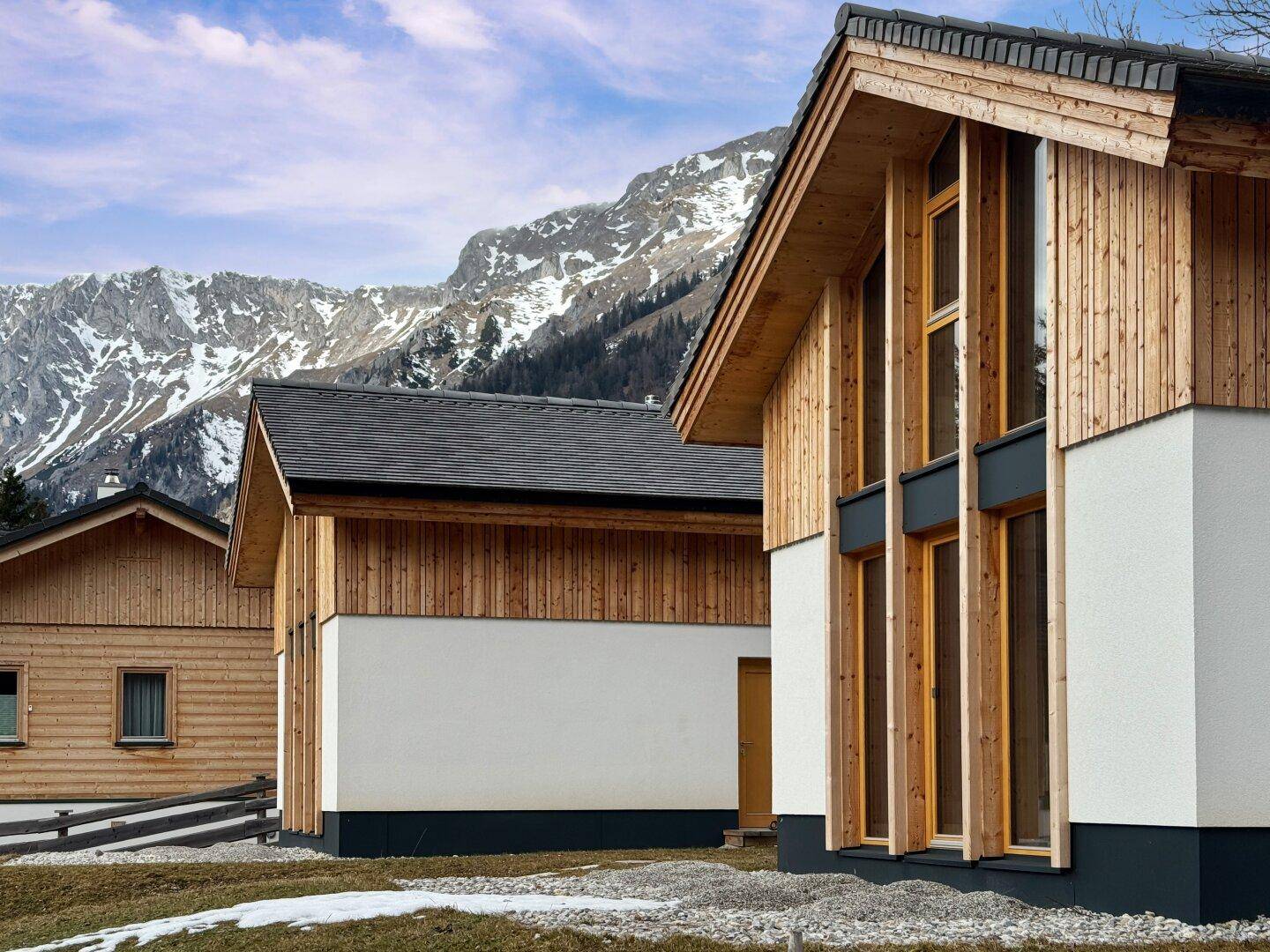 Seitenansicht eines modernen Chalets mit Holzverkleidung und weißer Fassade vor alpiner Kulisse.