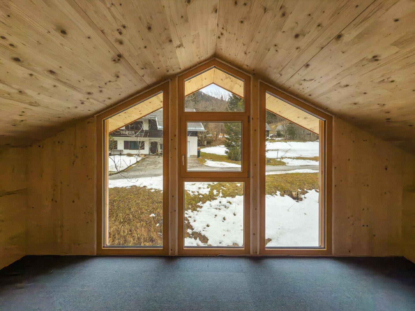 Obergeschossraum mit Holzdecke, großen Giebelfenstern und weitem Blick auf die verschneite Umgebung.