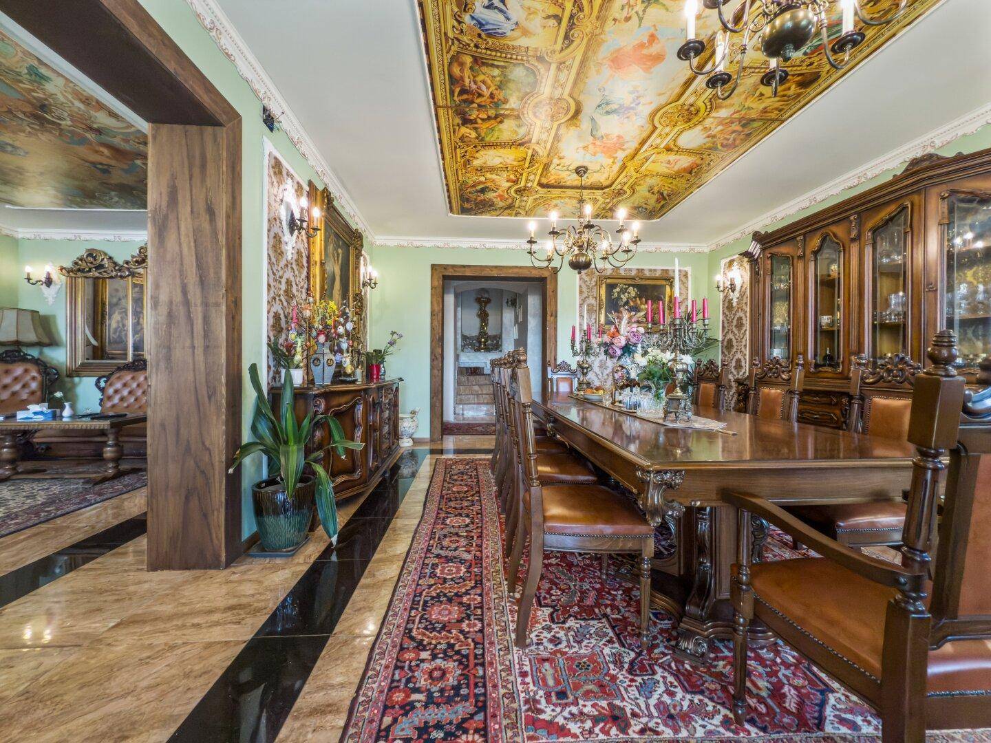 Prunkvolles Esszimmer mit großem Holztisch, verzierten Stühlen und einer kunstvoll bemalten Decke.
