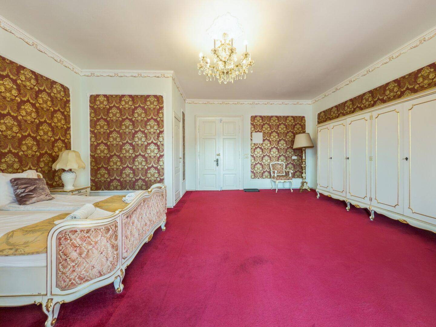 Großes Schlafzimmer mit opulentem Bett, weißen Einbauschränken und rotem Teppichboden.