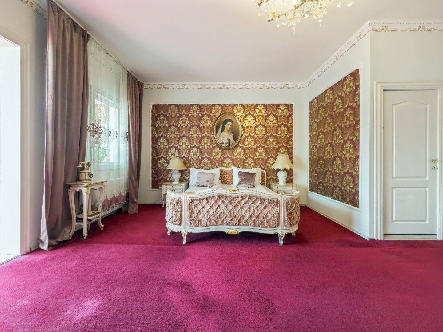 Elegantes Schlafzimmer mit einem verzierten Bett, gemusterten Tapeten und rotem Teppichboden.