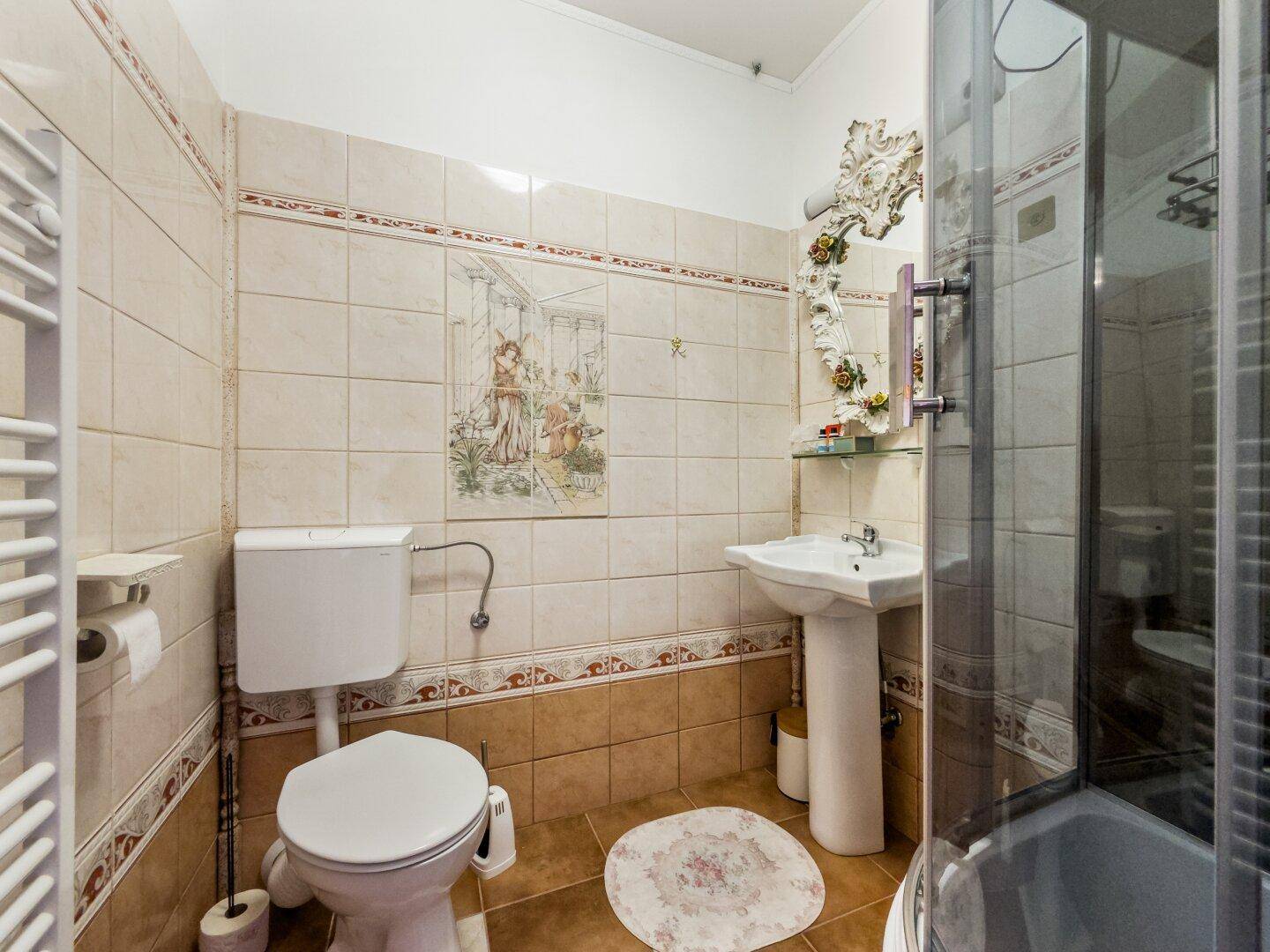 Badezimmer mit beigefarbenen Fliesen, Duschkabine, WC und Waschtisch mit verspiegeltem Aufsatz.