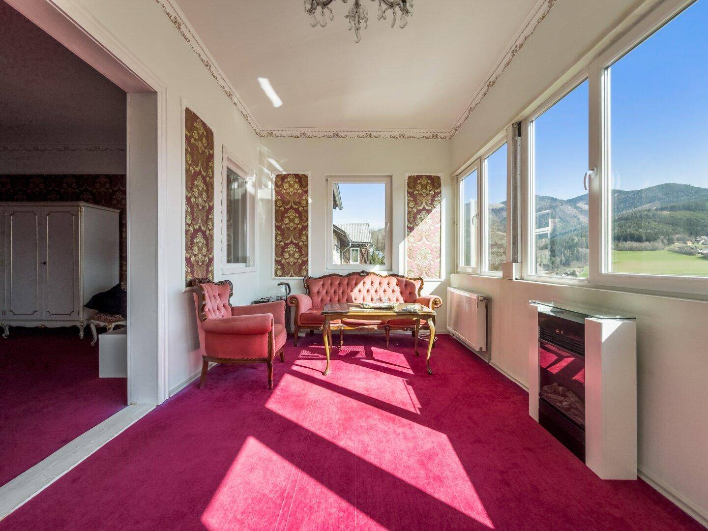 Helles Wohnzimmer mit großen Fenstern, rosafarbenem Teppichboden und Bergblick.