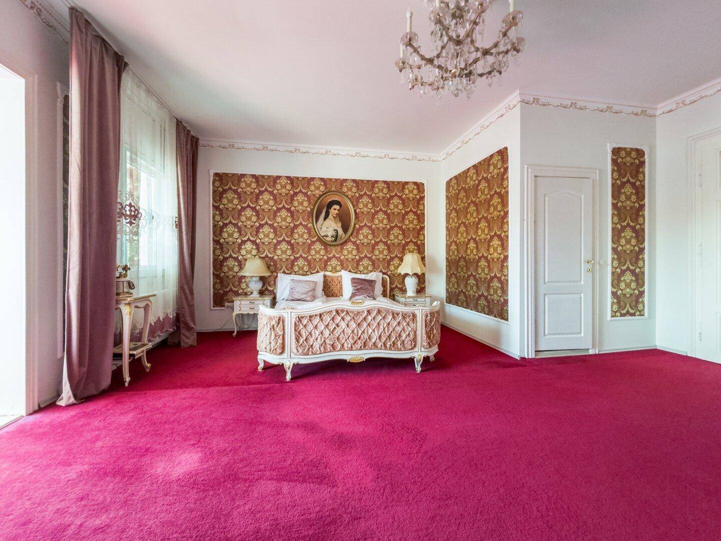 Großes Schlafzimmer mit opulentem Bett, rosafarbenem Teppichboden und gemusterter Tapete.