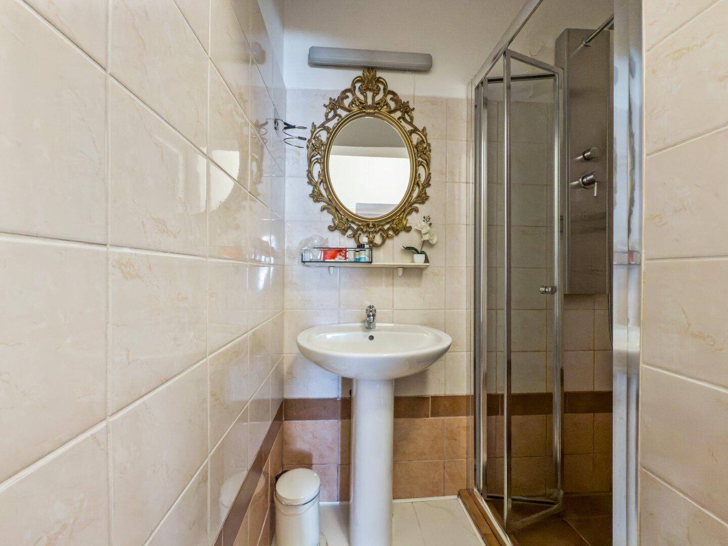 Badezimmer mit Duschkabine, Waschtisch und einem ovalen, reich verzierten Spiegel.