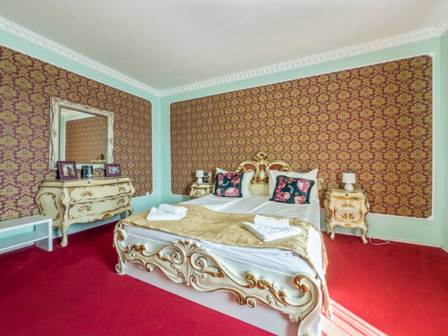 Schlafzimmer mit großem Bett, rosafarbenem Teppichboden und auffälliger Tapete in Goldtönen.