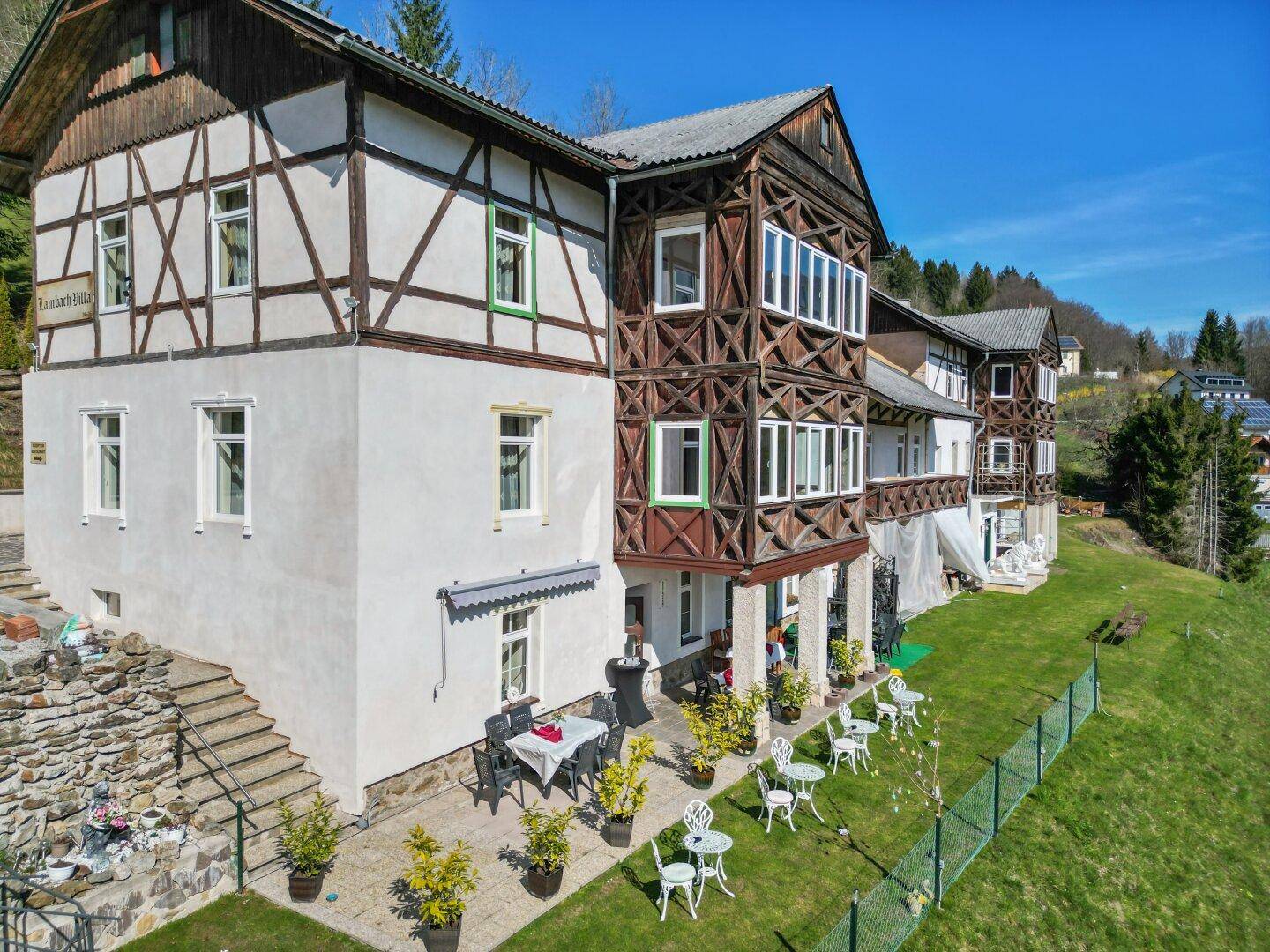 Traditionelles Gebäude mit Holzbalkonen und einer möblierten Terrasse, eingebettet in eine grüne Hügellandschaft.