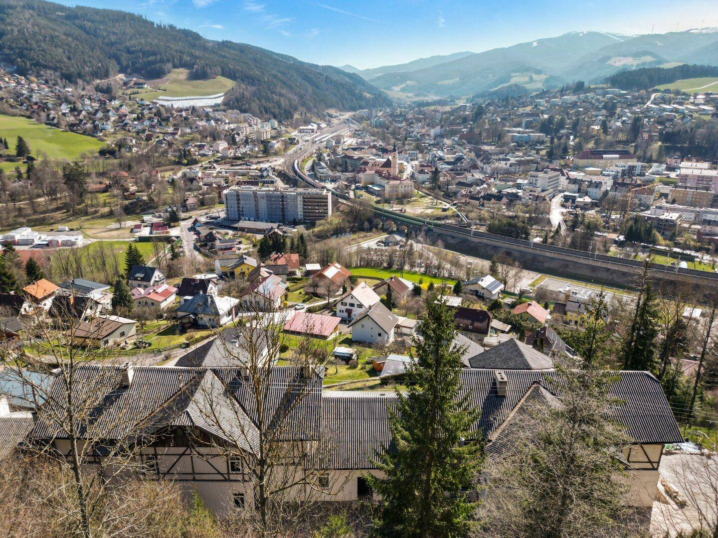 Panoramablick über eine malerische Stadt im Tal, umgeben von bewaldeten Bergen unter blauem Himmel.