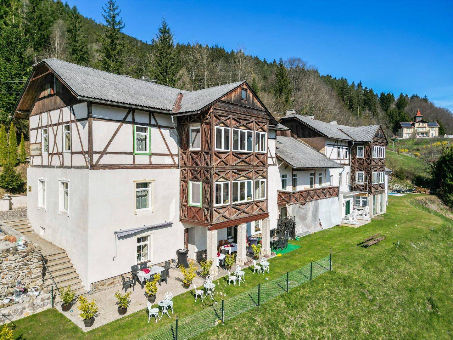 Längliche Gebäudefassade mit traditionellen Holzelementen und mehreren Terrassenbereichen in ländlicher Umgebung.