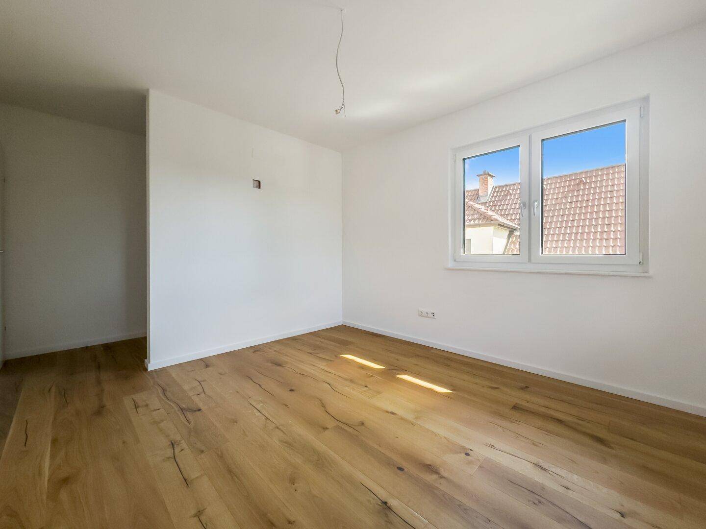Unmöbliertes Schlafzimmer mit Holzboden und Fensterblick auf ein benachbartes Dach, hell und ruhig.