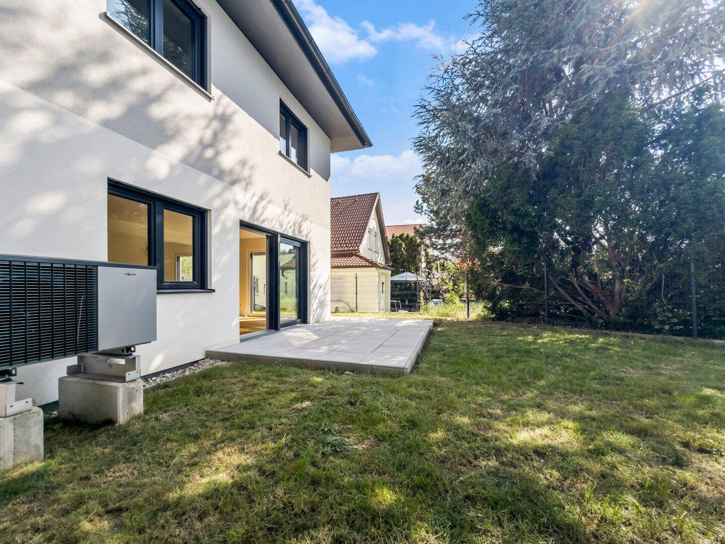 Seitlicher Blick auf das Haus mit Terrasse, Rasenfläche und einem Wärmepumpen-Außengerät im Garten.