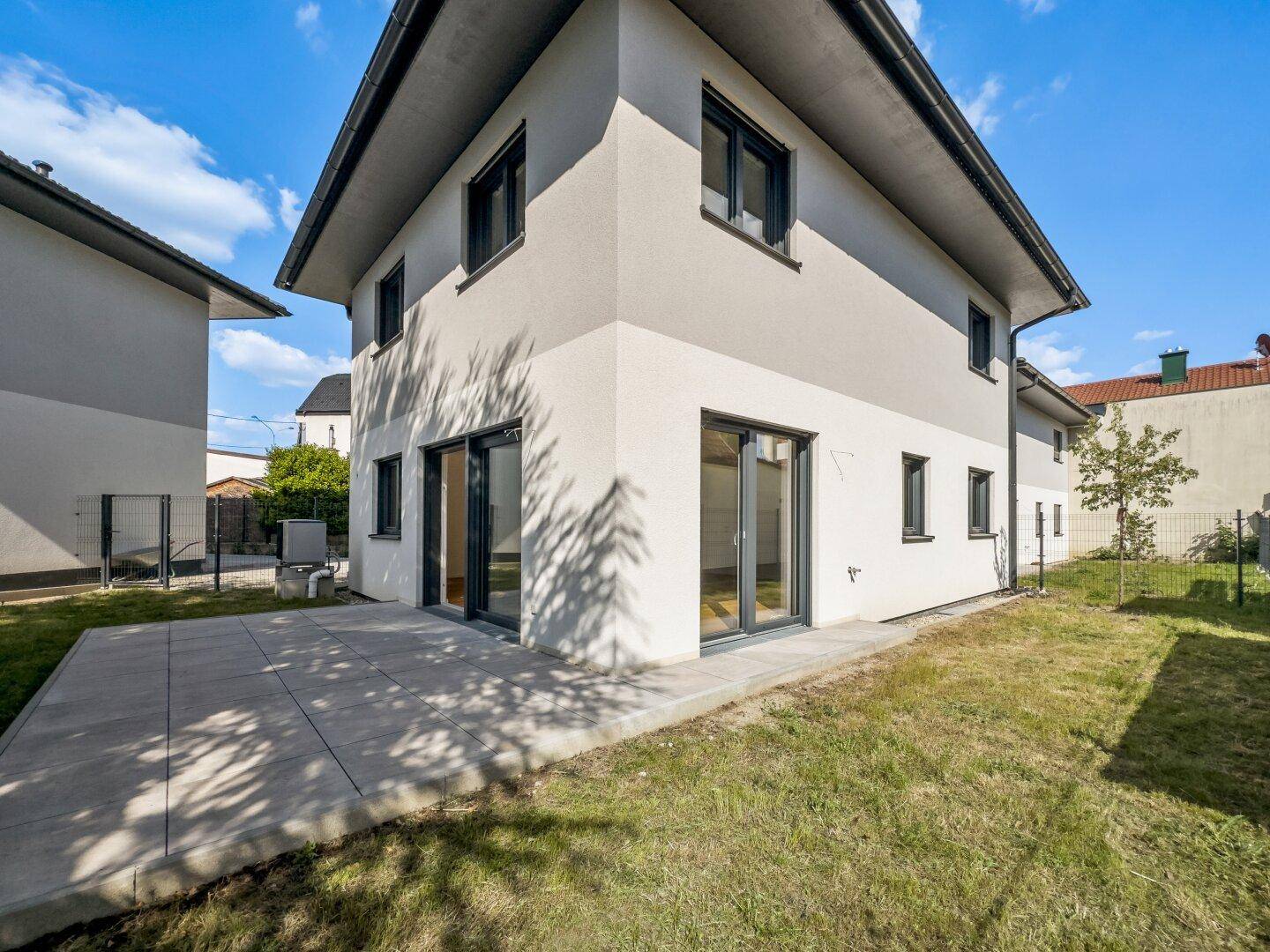 Neubau mit moderner Fassade, großer Terrasse und weitläufigem Gartenbereich unter blauem Himmel.