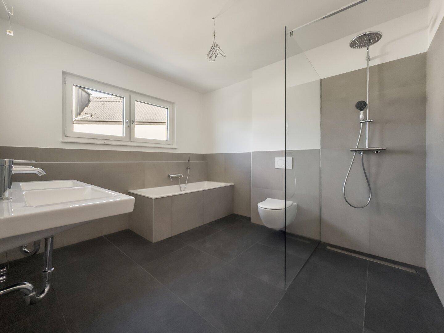 Modernes Badezimmer mit Badewanne, Dusche, Waschbecken und Fenster, in eleganten Grautönen gehalten.