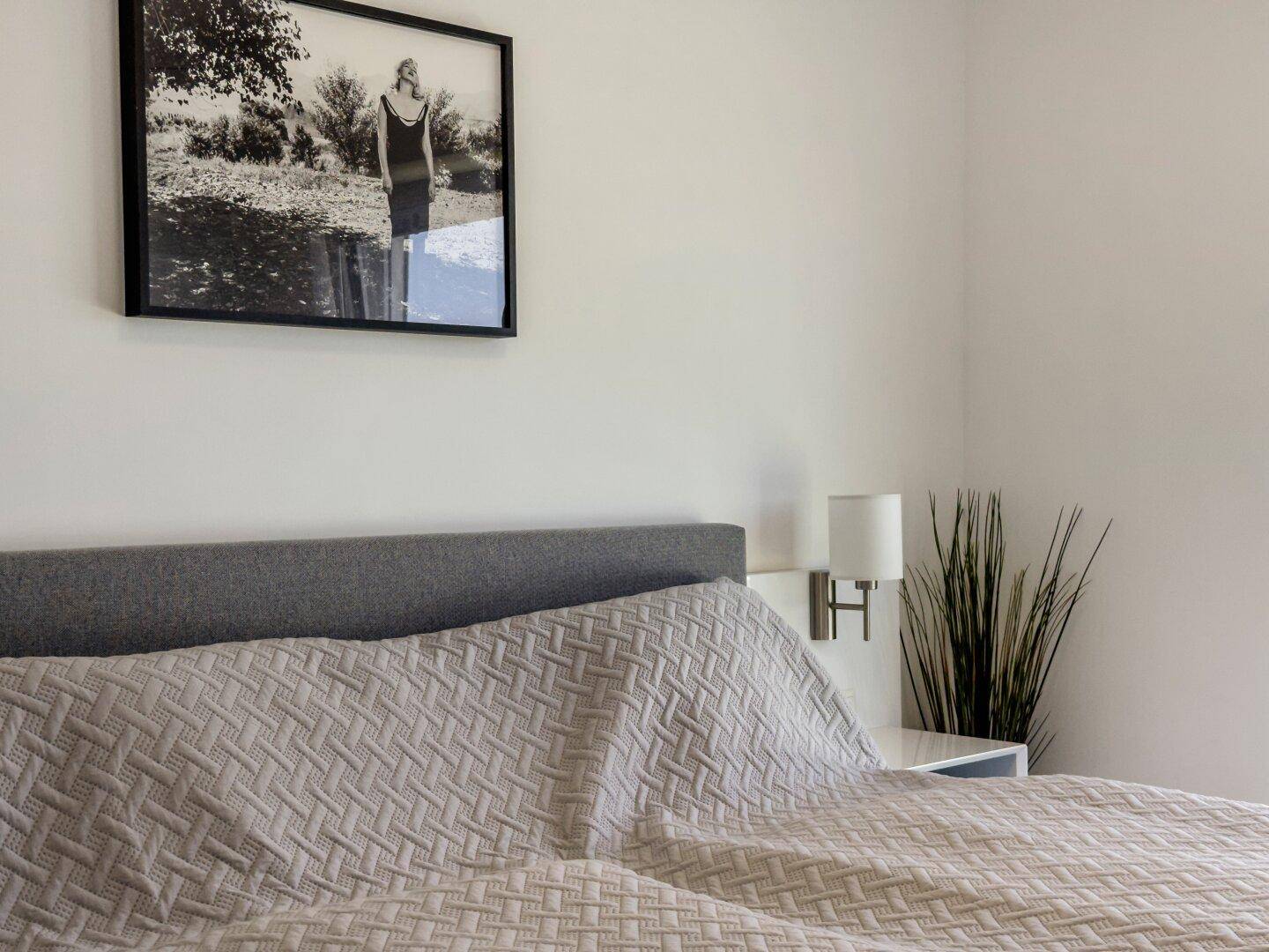 Detailansicht des Bettes im Schlafzimmer mit Wandleuchte und gerahmtem Bild.