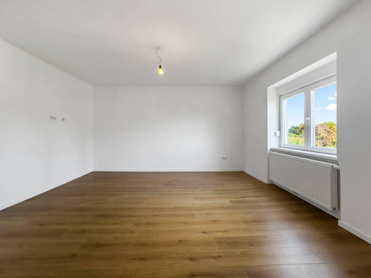 Helles, leeres Zimmer mit Laminatboden und Fenster, das viel Tageslicht hereinlässt.