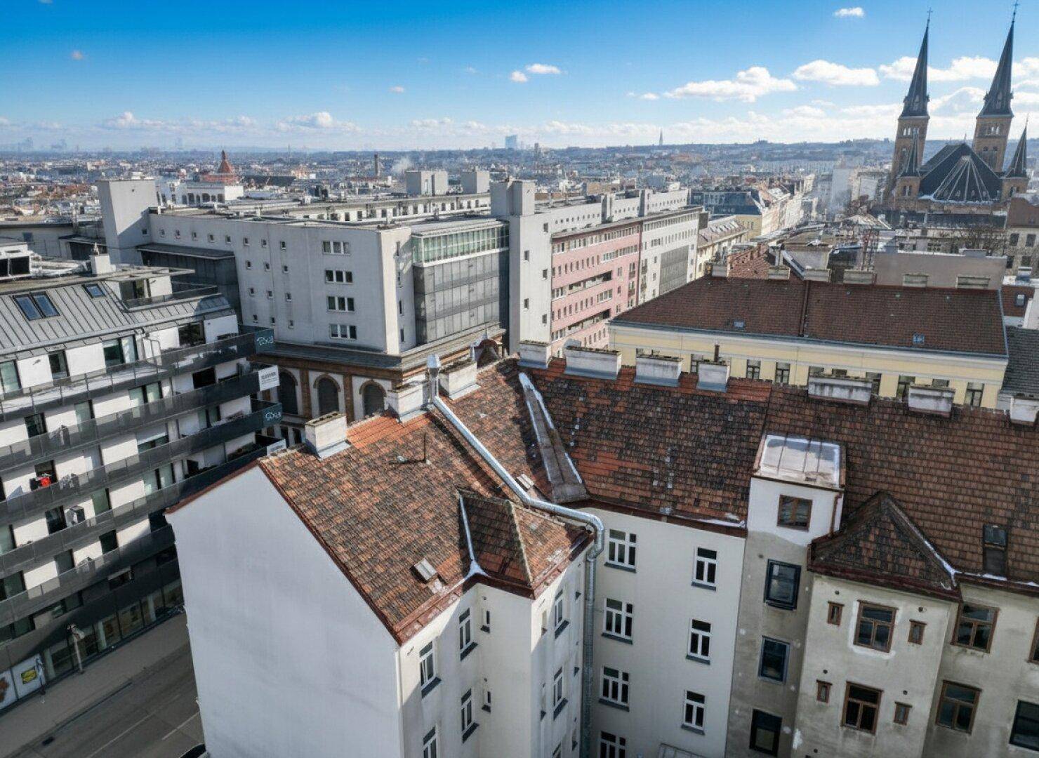 Luftaufnahme einer Stadtlandschaft mit verschiedenen Gebäuden und einer Kirche im Hintergrund unter blauem Himmel.