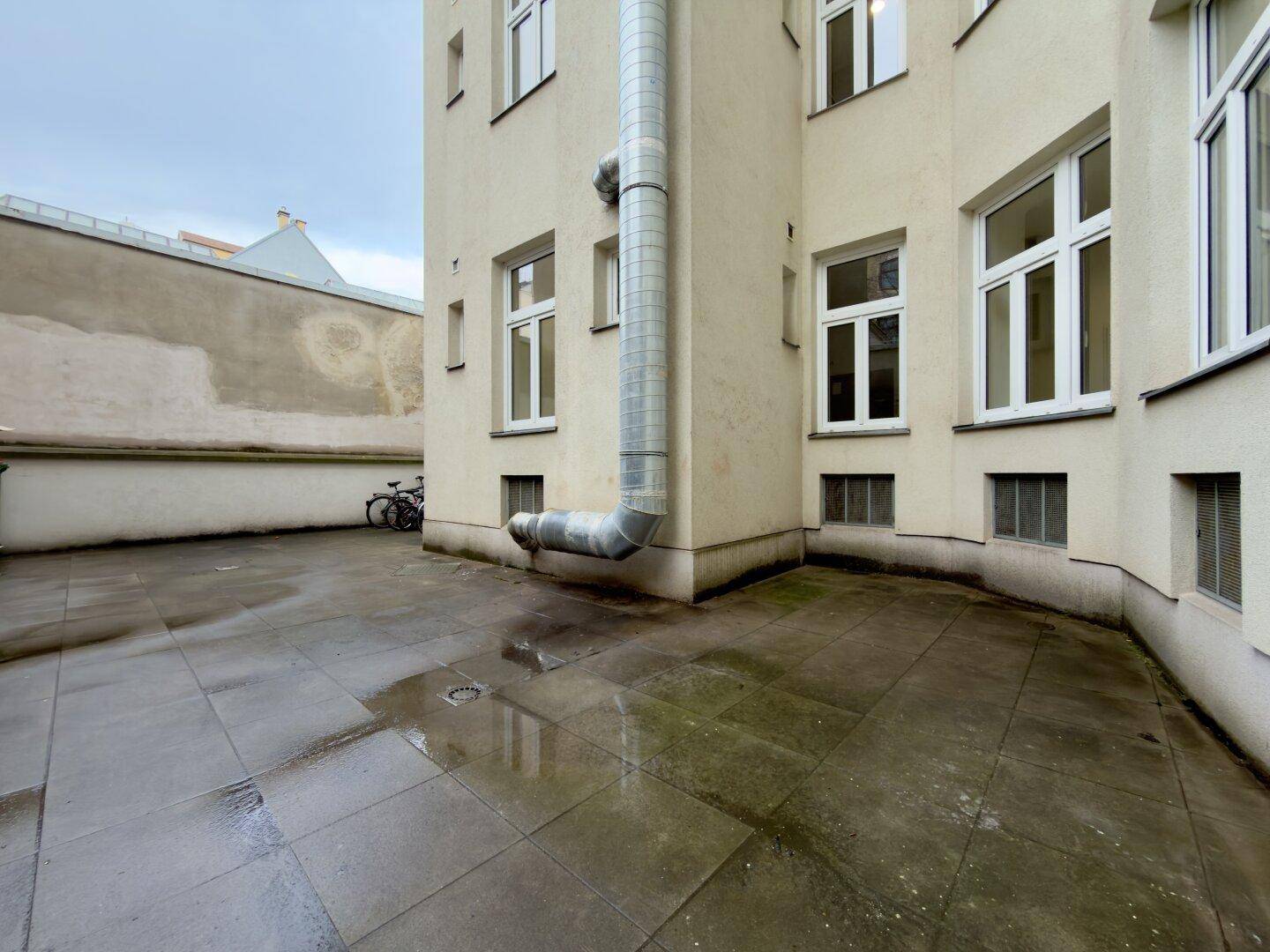 Gepflasterter Innenhof eines Gebäudes mit Entwässerungsrohr und Fenstern.
