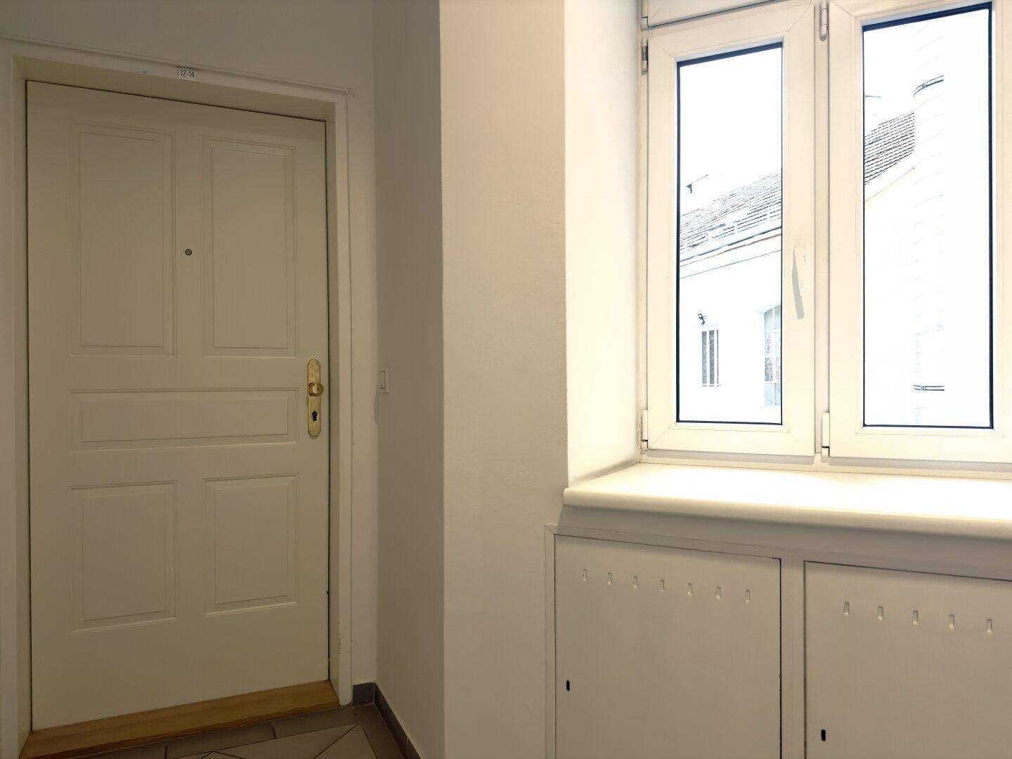 Heller Flur mit weißer Eingangstür, Fenster und Heizungsverkleidung.