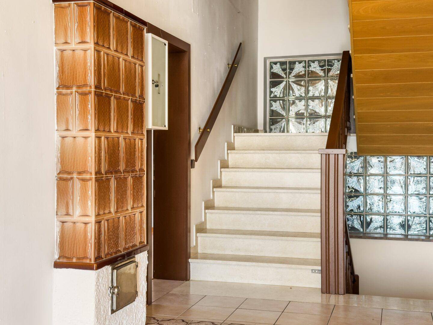 Treppenhaus mit hellem Fliesenboden, Kachelofen und Glasbausteinen für natürliches Licht.