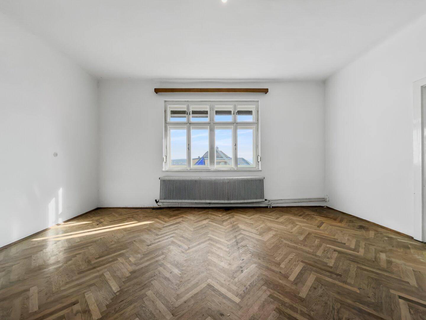Unmöbliertes Schlafzimmer mit Fischgrätparkett, weißen Wänden und großem Fenster, hell und geräumig.